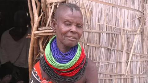 BBC A woman with colourful beads around her neck squints at the camera.