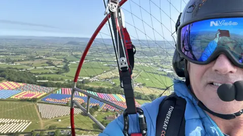A selfie of James Pearce paramotoring above Glastonbury Festival. He is wearing a blue puffer coat, reflective blue goggles and a black helmet. Behind him, you can see the harness and motor, which is strapped to him. In the distance, you can see the colourful tents being set up on Worthy Farm. 