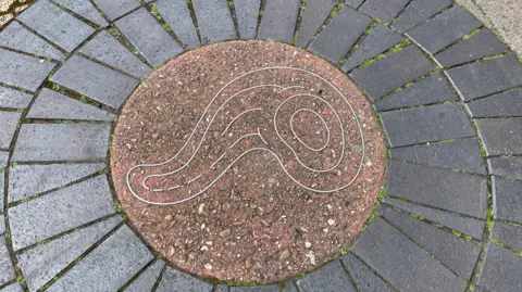 Alexa Rusakoff/BBC An image of a bacteria cell outlined in metal and set in stone, paved into the ground. The cell is curved in shape with a set of circles at one end and a long protruding shape on the left.