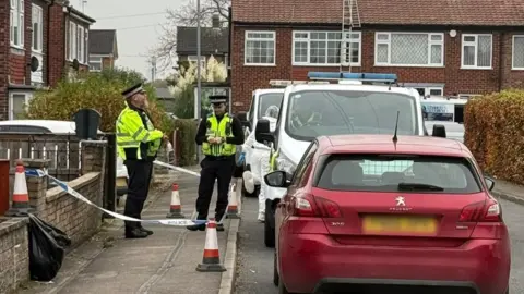 Two police officers stand outside a two-storey, terraced property which has been cordoned off with blue and white police tape. 