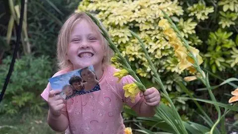 Nicola Hellem A child with blonde hair wears a pink top adorned with rainbows and a cheeky grin as she holds a photograph featuring her as a toddler, her father and her mother on a sunny day at the beach.