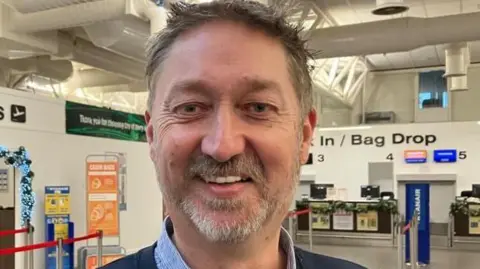 Steve Frazer has short brown hair and a beard.
He is standing in the City of Derry Airport main lounge.