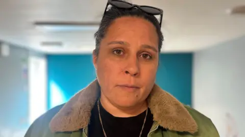 Nancy Magon is wearing a green jacket with brown fur on top, a black top and a gold chain and black sunglasses on top of her head. Her dark hair is tied back and she is looking at the camera.
