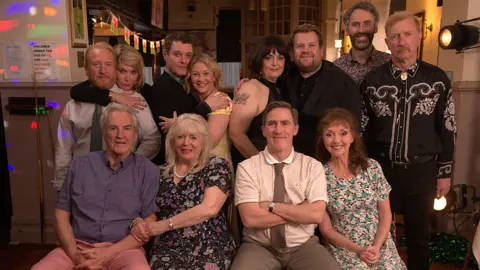 Cast line up in two rows to pose for a wedding celebration photograph but love's loser Dave Coaches doesn't appear happy