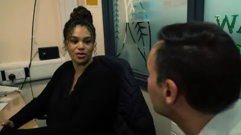 Gemma Ronald has black curly hair and is wearing a black long sleeved top. She is sat at an office desk and is talking to a man with dark hair.