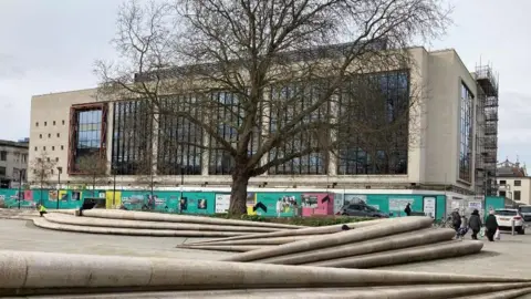 The new University of Gloucestershire campus in Gloucester city centre. It is concrete with large glass windows, bordered by green billboard fencing promoting the new campus. It is in Kings Square in Gloucester, with sweeping tiered concrete step-like structures through the square for people to sit on in the foreground. There is a large tree in front of the university campus.