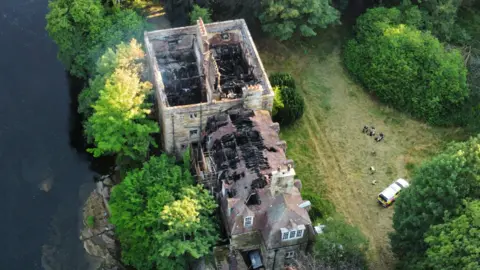 A drone image of Armathwaite Castle showing the damage caused by a fire. The building has a main rectangular structure with no roof and another structure connected to it, which is more similar to a house in shape. The roof is partially still on this section. Inside the affected areas are completely black with debris and collapsed beams and supporting structures. There is a river on one side. On the other side there is a lawn with an emergency vehicle parked on it and a group of people standing nearby.