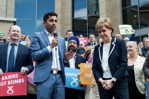 A large group of people stand outside a building during a public gathering, holding colourful signs with campaign slogans. One person speaks into a microphone while another stands beside them, with others holding placards in the background.