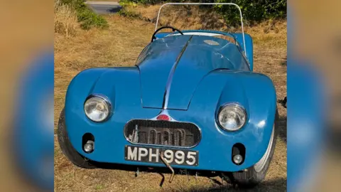 Stacey's Auctioneers A blue Connaught L2 sports car, which has large headlights and is parked on grass. Its engine is exposed thanks to a mesh grille and it has a long bonnet. The small car has two-doors.