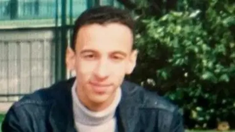 Family handout Hassan Bousanguar has short black hair and is wearing a black jacket and a cream roll-neck jumper. He is smiling outdoors in front of a bush and fence.