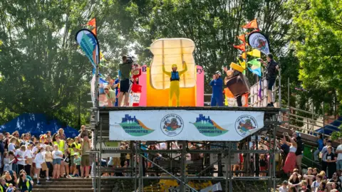 Bristol Picture A man wearing a costume in the shape of a piece of toast. He is standing at the edge of a platform supported by scaffolding. Crowds surround the platform and it is a sunny day. 