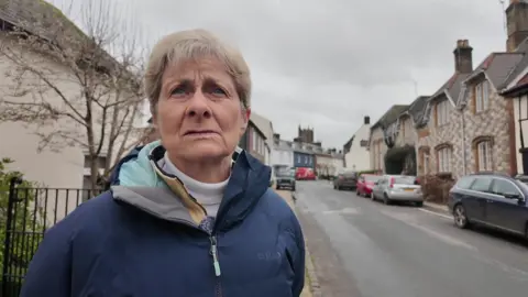 Helen Brown, a woman with short hair wearing a blue coat, stands on a pavement in the village of Cerne Abbas.
