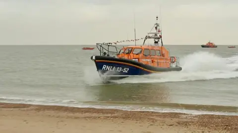 A new lifeboat is speeding through the water at Clacton-on-Sea. It is orange and blue and bears the name RNLI 13-52 painted in white on the side. Sea spray can be seen as it sails through the water