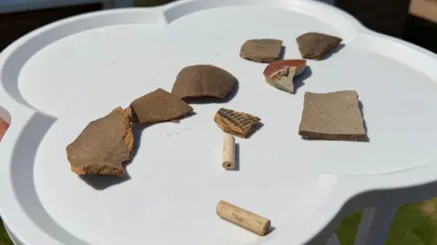 Becki Bowden / BBC Shards of pottery and other material on a white, clover-shaped table. One of the shards has a black pattern on it, the others are plain and brown in colour.