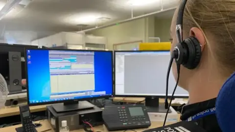 A woman in uniform with a headset looks at two computer screens.