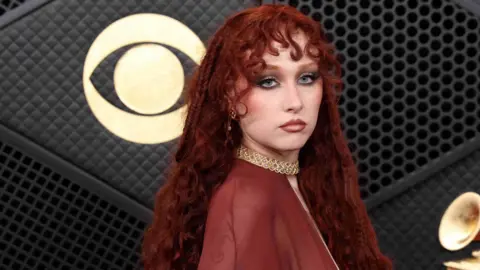 Chappell Roan at the Grammy Awards in a red dress. Her hair is long and curly. The background is black with a golden eye-style symbol.  