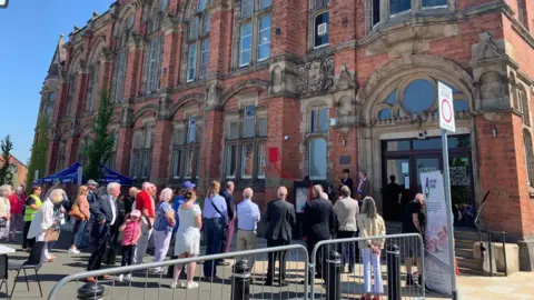 David Unwin A group of about 20 people are gathered outside a large historic red brick building were a small red cloth is covering a plaque on the wall