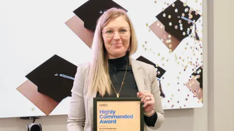 NHBC A woman with long blonde hair and glasses holds a framed certificate. She is wearing a black top, gold jewellery and a cream blazer.