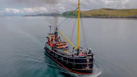 The Clyde Puffer SL VIC 32 - a small steamboat in dark blue/black, with a yellow mast, sails through water.