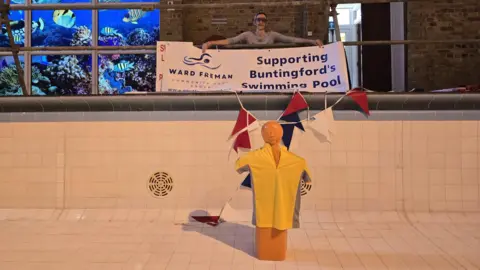 Ward Freman Community Pool Group A woman inside an empty swimming pool holding a banner supporting Buntingford’s community pool, with a yellow mannequin in a life jacket in the foreground. There is bunting in the pool and a poster, that looks like a fish tank to the left. The pool is made of cream tiles. 