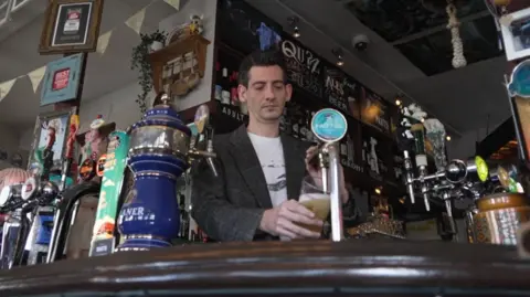 A dark-haired man pulls a pint with a bar in the background. The picture is taken from a low angle