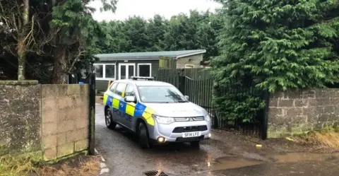 A police car sits at the entrance of a kennel, with green buildings in the background