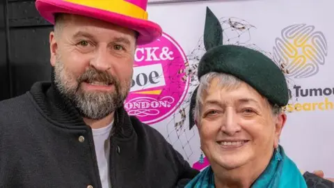 Alfie Boe and Anne McEntee. He is wearing a pink top hat and she is wearing a blue scarf and green hat