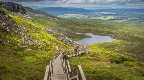 Getty Images The Cuilcagh Boardwalk Trail - a very long, very steep wooden staircase which crosses an expansive bog in County Fermanagh. The camera is pointing down the staircase which stretches into the distance across open land. A lake is partially visible in the middle of the photo. 