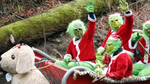 Sam Hepburn People dressed in green Grinch masks and red santa outfits waving towards the camera from the top of a raft