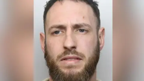 A mugshot of a man with blue eyes, a moustache and beard