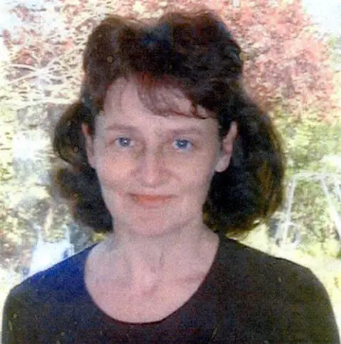 Wiltshire Police A grainy photograph of a woman with shoulder-length brown hair. She is wearing a black scoop-neck top and is standing in front of a tree smiling.