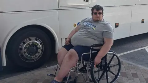 Charley Jonstone-Brent Charley Jonstone-Brent who uses a mobility aid, sitting in a wheelchair in front of a coach. It's a bright day and he is wearing sunglasses, shorts and a t-shirt.