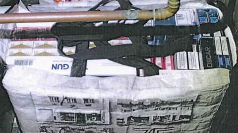 LDRS A close-up image shows a black rifle partially concealed inside a white reusable shopping bag. The bag has a printed black-and-white illustration of buildings and a handle looped over the firearm. The visible portion of the weapon includes a grip, trigger area, and part of the barrel extending across the top of the image. Surrounding the bag are stacks of boxed items arranged horizontally and vertically, including cartons with visible labels such as “GUN” and “OSC...,” suggesting packaged goods or supplies. The items appear tightly packed in a confined storage space, with muted lighting and a slightly grainy image quality.