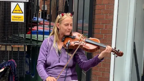 Grace Wood/BBC A woman with long blonde hair, wearing a purple fleece plays viola. She is standing on a street corner and wearing a headband with cat ears.