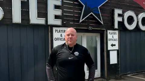 Gareth Edwards stands looking at the camera in front of the football club entrance. He has short white hair and is wearing the Newcastle Blue Star FC kit.