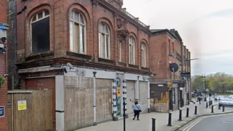 Google A derelict red brick building is boarded up on the ground floor. The first floor has arched windows which are partially boarded up. A woman wearing black trousers and a blue jacket walks past the building.
