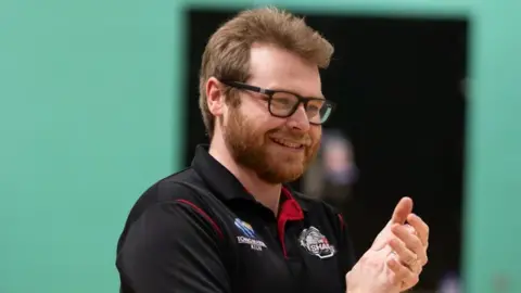 Solent Sharks Ricky Goddard clapping and standing in front of a green background. He is wearing a black Solent Sharks T-shirt and has a brown beard, brown hair and dark-rimmed glasses.