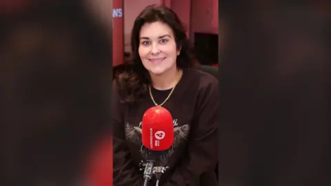 Woman with long dark hair, wearing a brown sweatshirt and a gold necklace.