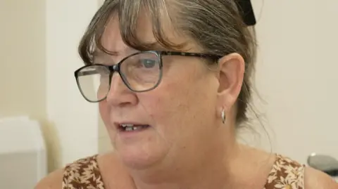 A head and shoulders view of Angela, wearing glasses with tied-back greying hair and hoop earrings