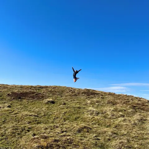 Eilidh McCallister A woman jumps on the top of a hill. The grass in the foreground is green. The woman jumping in the middle is wearing dark clothing. The sky is blue.