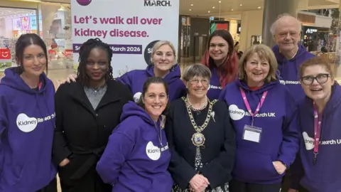 Kidney Research UK A group of eight women and one man standing in Queensgate Shopping Centre beside a sign that says "Let's walk all over kidney disease". Six of the women and the man are wearing purple Kidney Research UK hoodies. The seventh woman who is in the middle of the group, is wearing a mayoral chain over her black jacket. The eighth woman, second on the left, is wearing a black coat over a grey top.