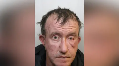 Adam Pugh looks at the camera for a mugshot. He wears a black hoodie, has thin black hair and red patches on his face