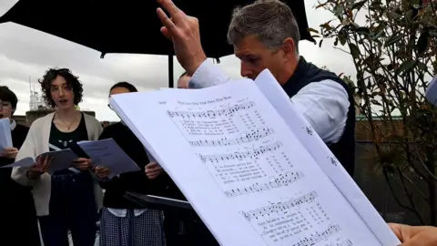 A sheet of music is in the foreground, with singers and a conductor seen beyond it.