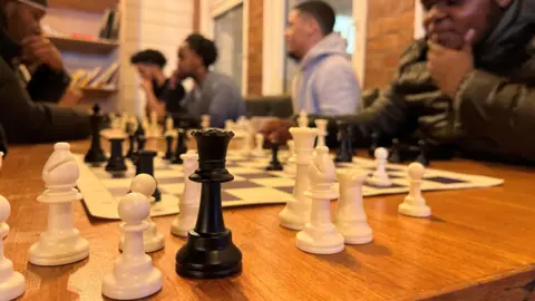 Chess pieces adorn the table nearest the camera with people playing the game in the background.