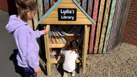 Sean Lawless Lydia Lawless is holding open the door of her mini library so her little sister can peruse the books. 