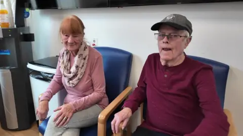 A wife and husband sit in chairs in a medical waiting room. There is a water cooler to their left