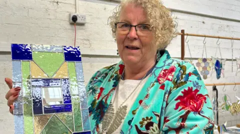 A lady with short blonde curly hair and glasses is smiling and holding up her stained glass art work. The art work has light green, light blue and dark blue square and triangle shapes making a collage. The lady is wearing a bright blue jacket with flowers on. Behind her is a white brick wall and some of her other stained glass art hanging up on a rail.