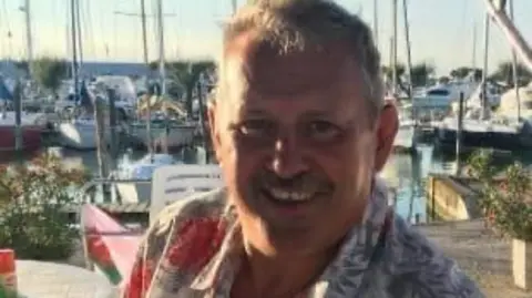 Mike O'Leary wears a tropical shirt, smiling in the sunshine at a harbour, boats visible in the background