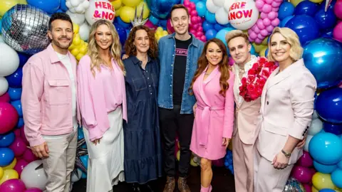 A line-up of three men and four women smiling at the camera for a promotional image, in front of a wall of pink, yellow, white and blue balloons.