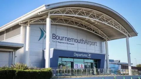 PA Exterior view of Bournemouth Airport departures terminal on a sunny day, featuring a large curved metal canopy supported by white columns, glass entrance doors, and prominent “Bournemouth Airport” and “Departures” signage against a clear blue sky.